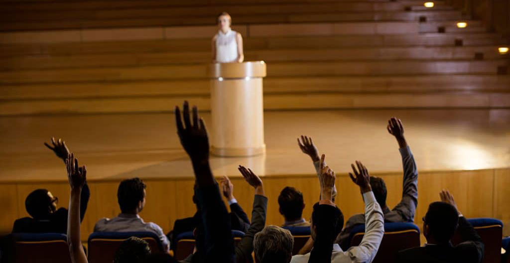 Assemblée générale de sociétaires d'une banque coopérative votant à main levée, salle de conférence avec intervenant au pupitre et écran affichant l'ordre du jour